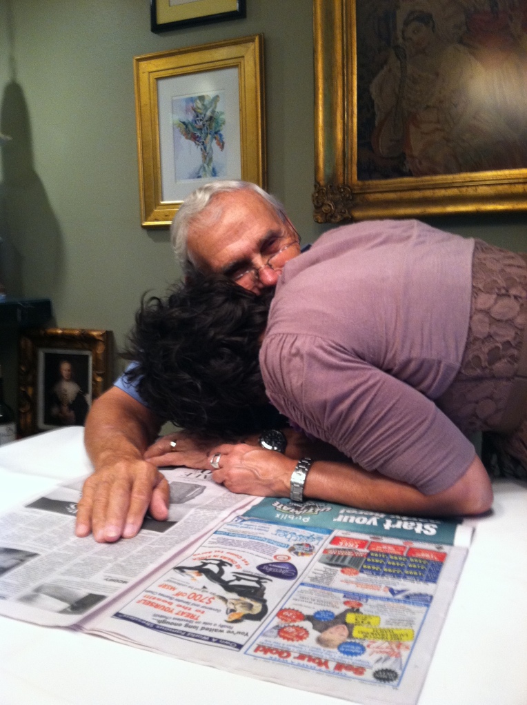 Jill leaning over her Dad to kiss him while he is reading the newspaper at his dining room table